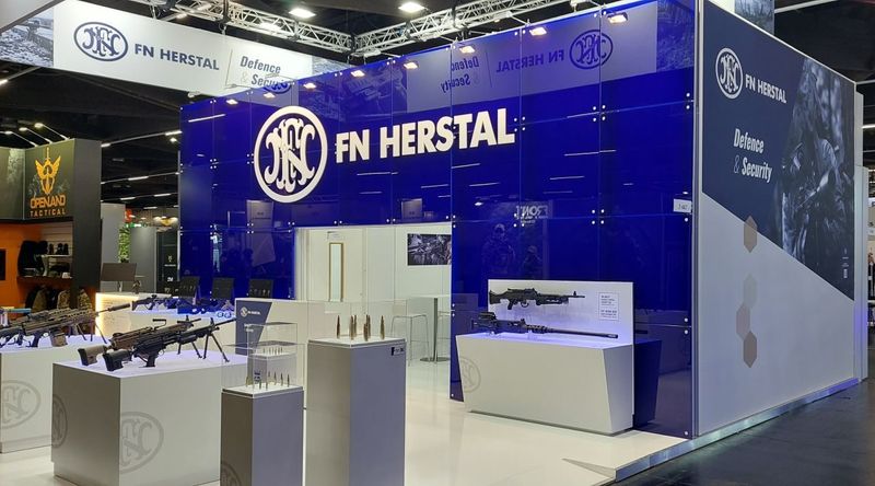 Stand van FN Herstal met een blauwe glazen wand en witte vitrines